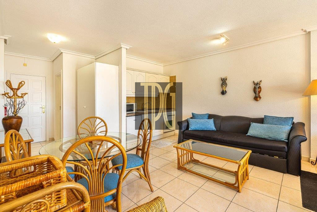 Living room with a round glass dining table and wicker chairs, adjacent to a dark sofa with blue pillows on a tiled floor.