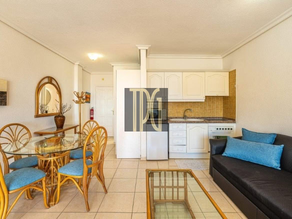 Open-plan living area with a glass dining table and bamboo chairs, a dark sofa with blue cushions, and a compact white-kitchen backdrop.