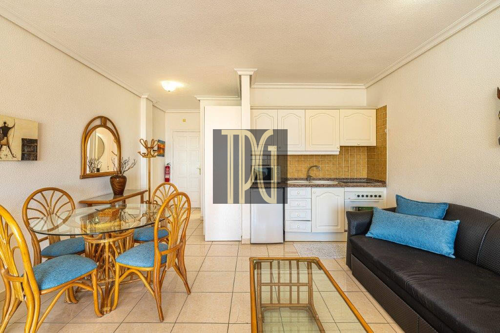 Open-plan living area with a glass dining table and bamboo chairs, a dark sofa with blue cushions, and a compact white-kitchen backdrop.