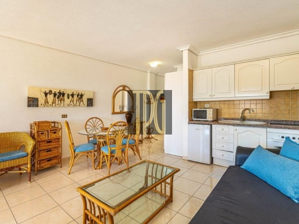 Open-plan living area with wicker seating, glass coffee table, and a blue-cushioned sofa facing a white-cabinet kitchen with beige tile backsplash.