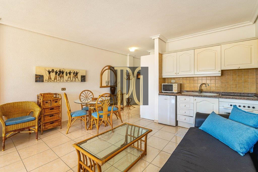 Open-plan living area with wicker seating, glass coffee table, and a blue-cushioned sofa facing a white-cabinet kitchen with beige tile backsplash.