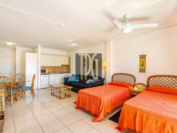 Suite-style hotel room with two orange-draped beds, wicker headboards, and a nightstand between them; a black sofa, glass coffee table, and a floor lamp nearby. An open kitchenette with white cabinets and a dining area with wicker chairs and blue cushions is visible, plus a ceiling fan above.