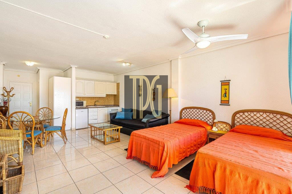 Suite-style hotel room with two orange-draped beds, wicker headboards, and a nightstand between them; a black sofa, glass coffee table, and a floor lamp nearby. An open kitchenette with white cabinets and a dining area with wicker chairs and blue cushions is visible, plus a ceiling fan above.