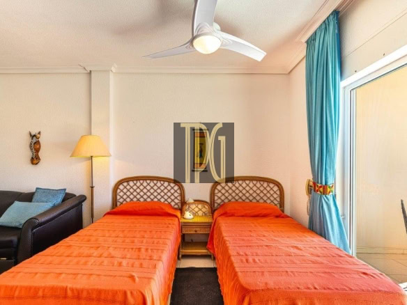 Hotel room with two twin beds in orange duvets, wicker headboard, and a seating area by a glass door with blue curtains.