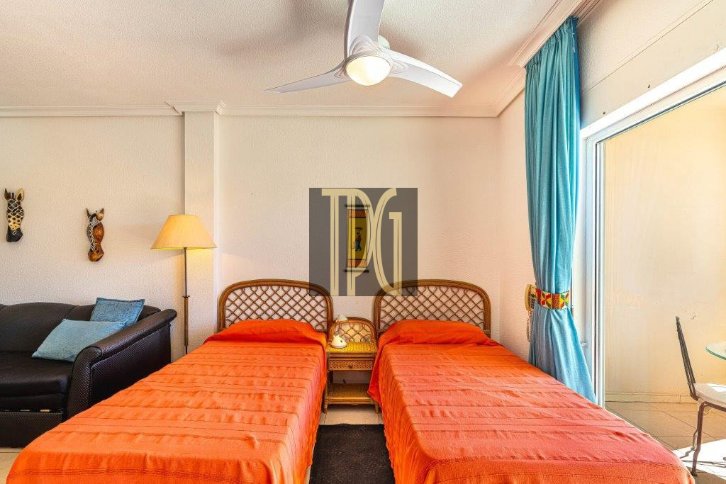 Hotel room with two twin beds in orange duvets, wicker headboard, and a seating area by a glass door with blue curtains.