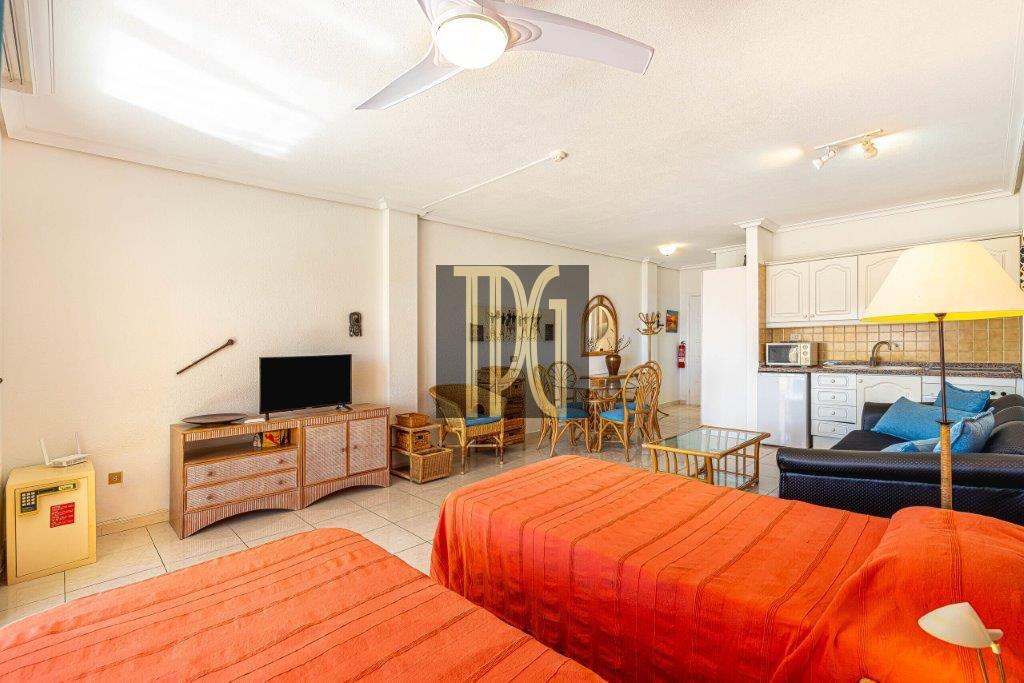 Spacious studio with two orange bedspreads in the foreground, a TV stand, wicker seating, and an open kitchen to the right.