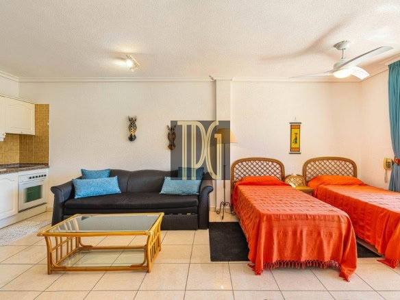 Hotel suite living area with kitchenette on the left, black sofa, glass coffee table, and two orange twin beds on the right.