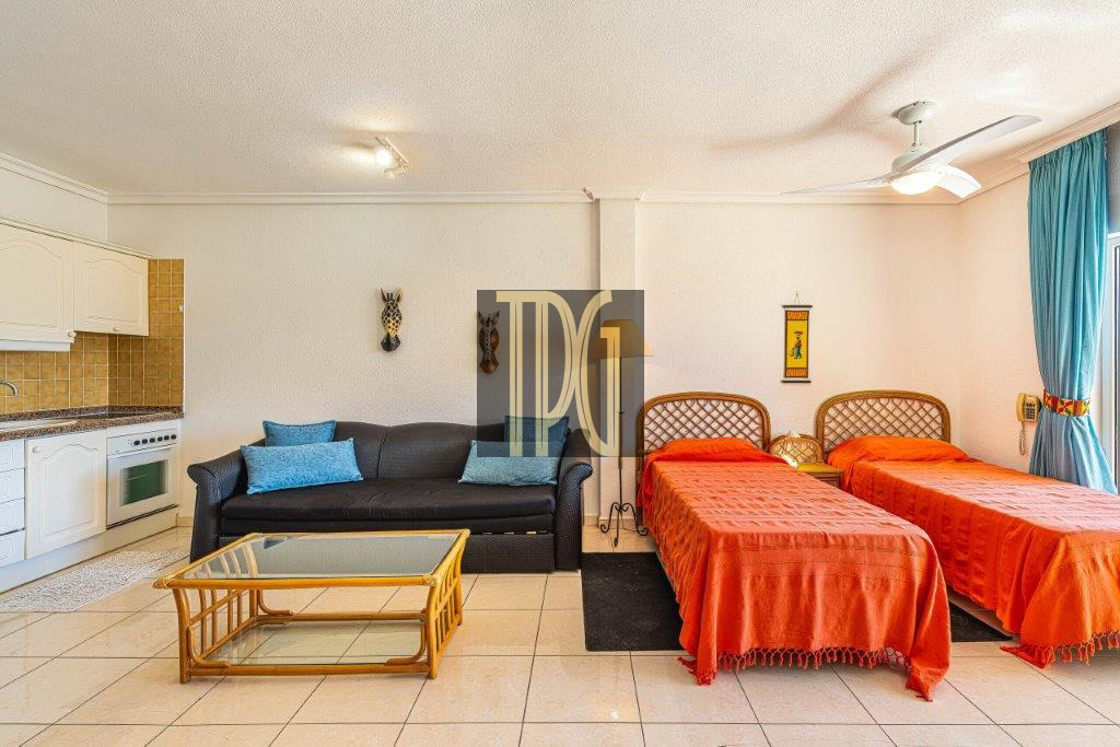 Hotel suite living area with kitchenette on the left, black sofa, glass coffee table, and two orange twin beds on the right.