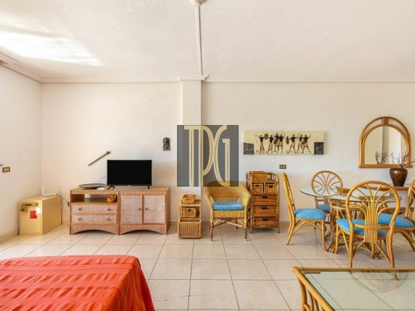 Bright open living/dining room with wicker furniture, a TV on a woven stand, and a round glass dining table with blue-cushioned chairs.