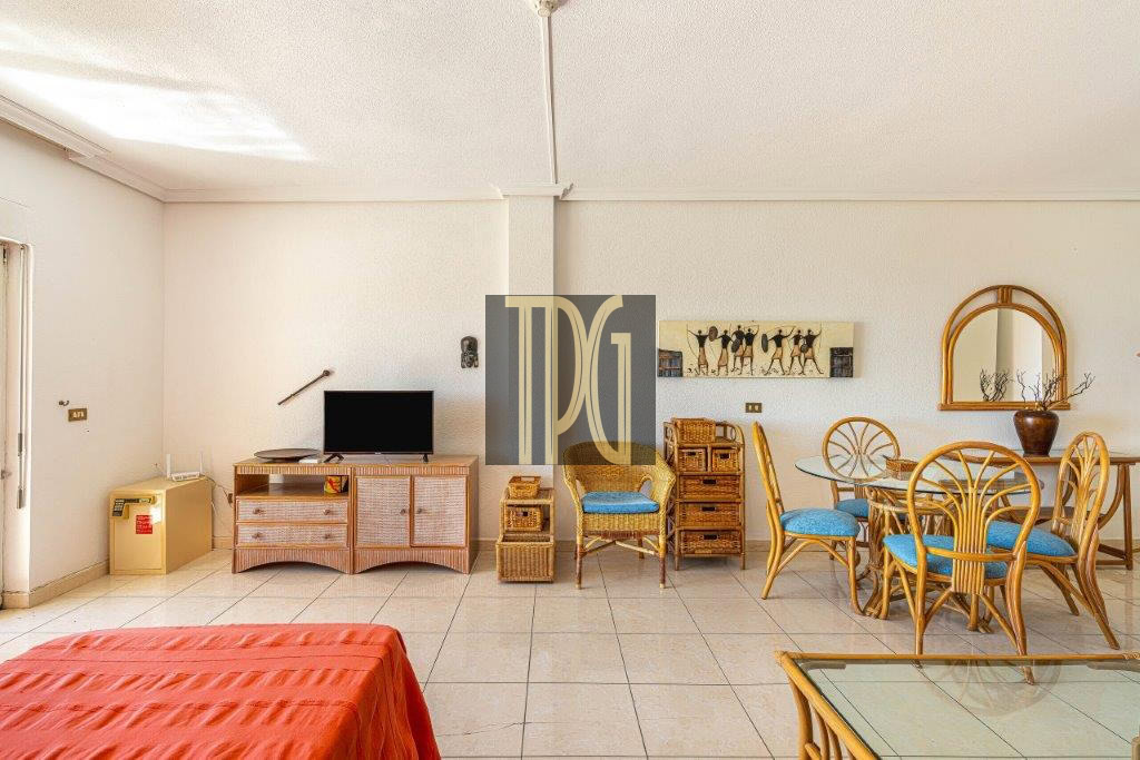 Bright open living/dining room with wicker furniture, a TV on a woven stand, and a round glass dining table with blue-cushioned chairs.