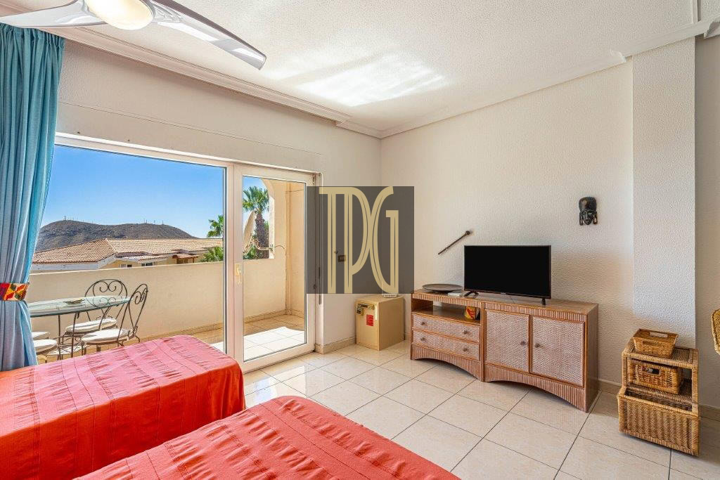 Twin-bed hotel room with red bedspreads, tile floor, and a wicker TV cabinet facing a flat-screen TV; sliding glass doors open to a small balcony with a table and chairs; blue curtains and ceiling fan above.