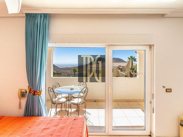 Hotel room interior with sliding glass doors to a balcony, turquoise curtain, orange bedspread, and a small round table with two chairs overlooking a coastal landscape with hills and sea.