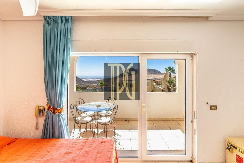 Hotel room interior with sliding glass doors to a balcony, turquoise curtain, orange bedspread, and a small round table with two chairs overlooking a coastal landscape with hills and sea.