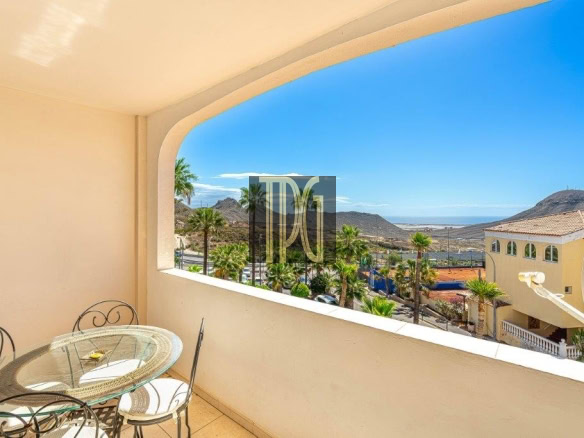 Balcony with a glass round table and wrought-iron chairs overlooking palm trees, hills, and a distant sea under a clear blue sky.