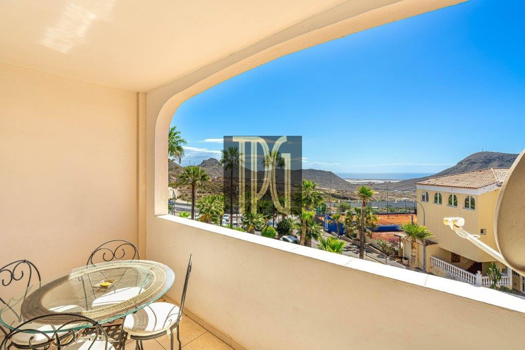 Balcony with a glass round table and wrought-iron chairs overlooking palm trees, hills, and a distant sea under a clear blue sky.