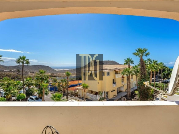View from a balcony overlooking a sunny coastal town with palm trees, a yellow apartment building, parked cars, and a satellite dish, with the sea in the distance.