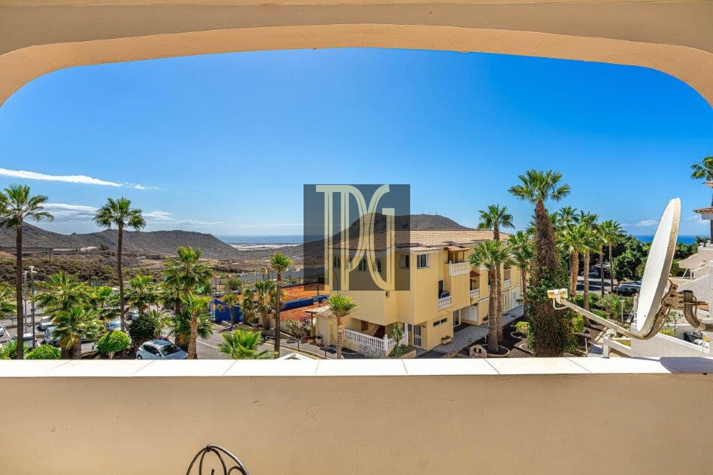 View from a balcony overlooking a sunny coastal town with palm trees, a yellow apartment building, parked cars, and a satellite dish, with the sea in the distance.
