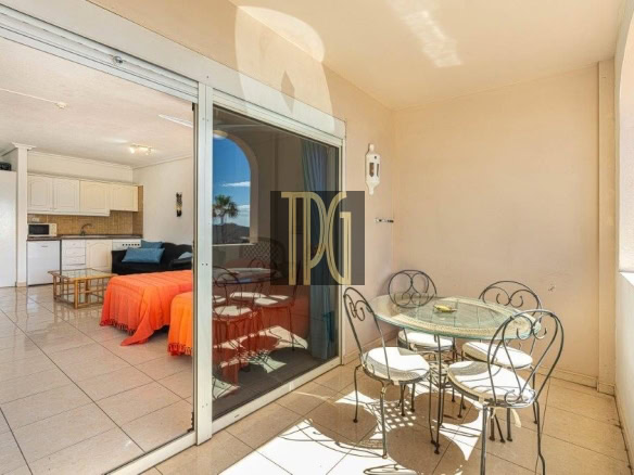 Balcony with a round glass table and four metal chairs; sliding glass doors reveal a bright open-plan living area with a kitchen and orange-covered seating.