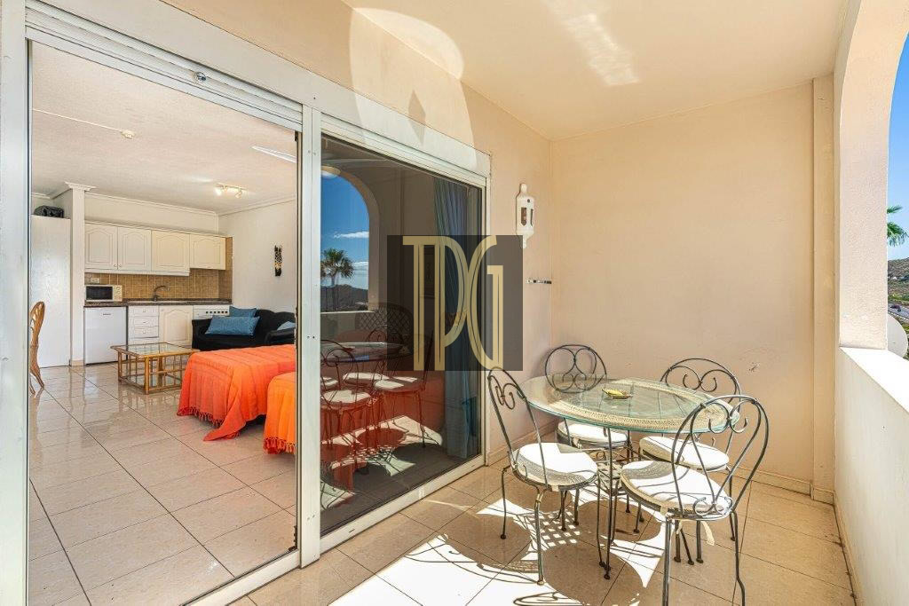 Balcony with a round glass table and four metal chairs; sliding glass doors reveal a bright open-plan living area with a kitchen and orange-covered seating.
