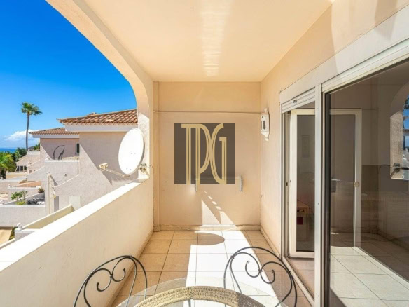 Sunlit balcony with tiled floor, round metal chairs, and a glass sliding door leading indoors; view of neighbouring buildings and blue ocean beyond.