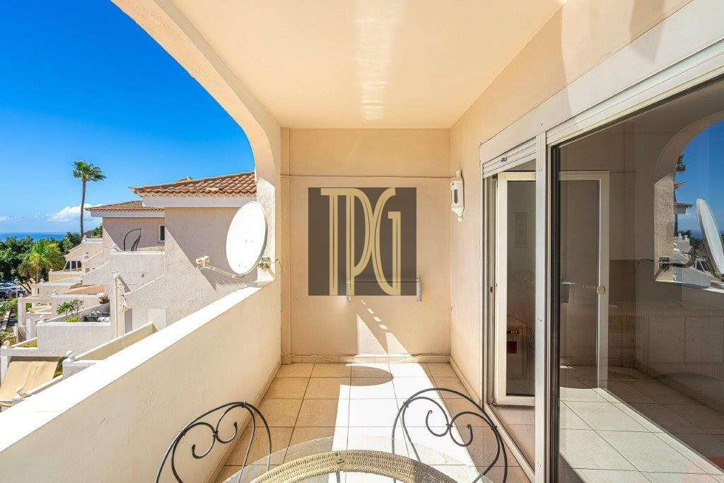 Sunlit balcony with tiled floor, round metal chairs, and a glass sliding door leading indoors; view of neighbouring buildings and blue ocean beyond.
