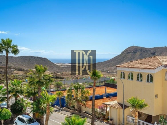 Sunny coastal scene with palm trees, a yellow villa, and a clay tennis court against distant hills and the sea.