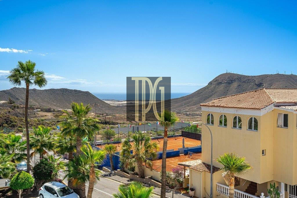 Sunny coastal scene with palm trees, a yellow villa, and a clay tennis court against distant hills and the sea.