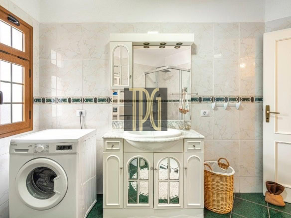 Bathroom with a front-loading washing machine beside a white vanity and arched mirror, tiled walls, and a wicker basket.