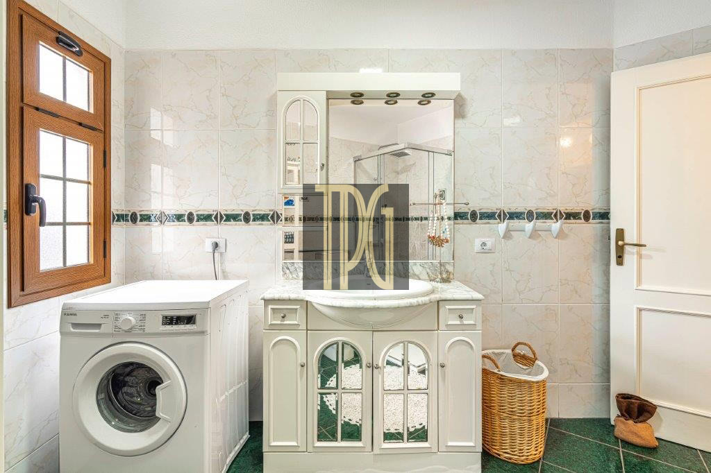Bathroom with a front-loading washing machine beside a white vanity and arched mirror, tiled walls, and a wicker basket.