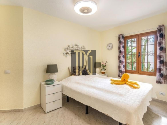Bright bedroom with a white bedspread, yellow walls, and a large window with patterned curtains; an orange towel folded like an octopus on the bed.