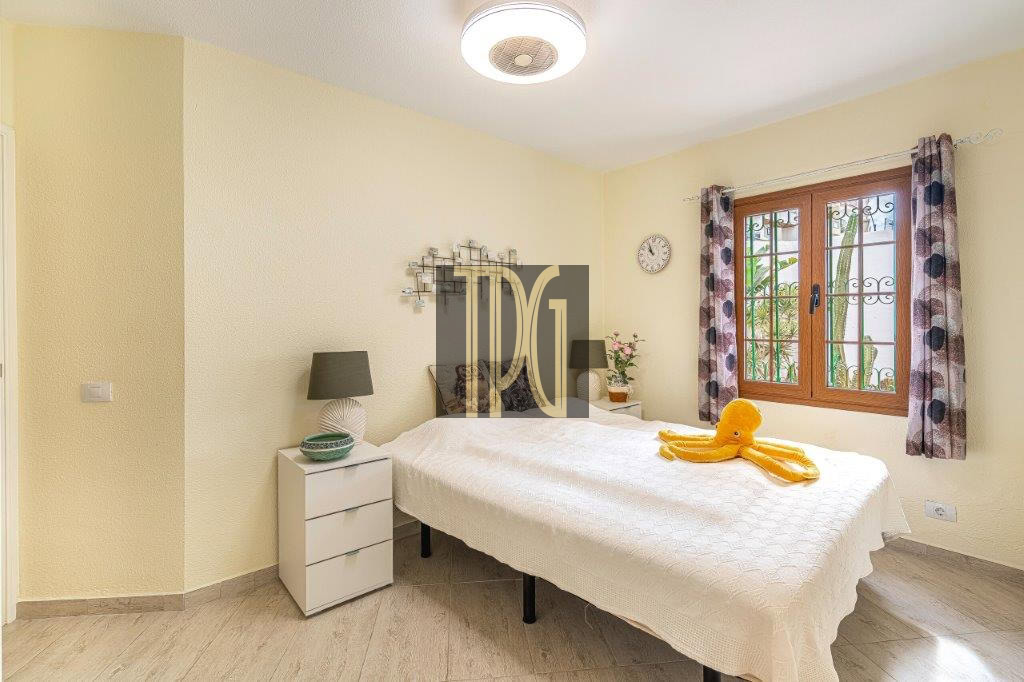 Bright bedroom with a white bedspread, yellow walls, and a large window with patterned curtains; an orange towel folded like an octopus on the bed.