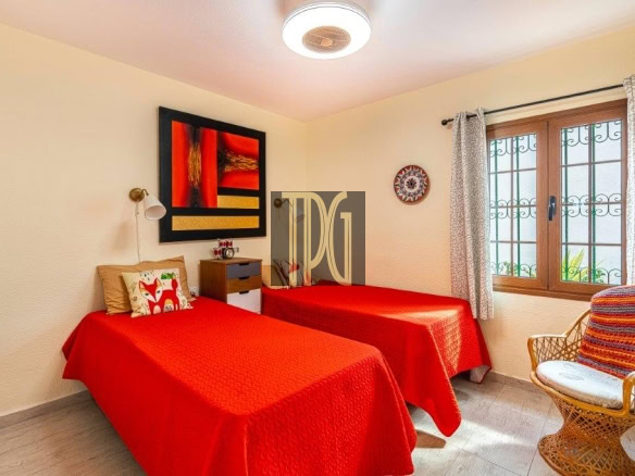 Twin bedroom with two single beds in red bedspreads, beige walls, and a wicker chair by a window with decorative bars.
