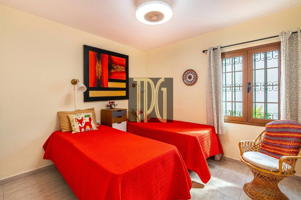 Twin bedroom with two single beds in red bedspreads, beige walls, and a wicker chair by a window with decorative bars.