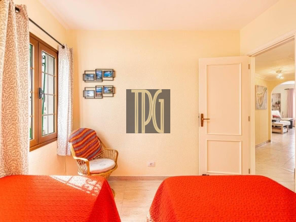 Sunlit bedroom with two red-covered beds, a wicker chair by a window, and an open door to an adjoining room.