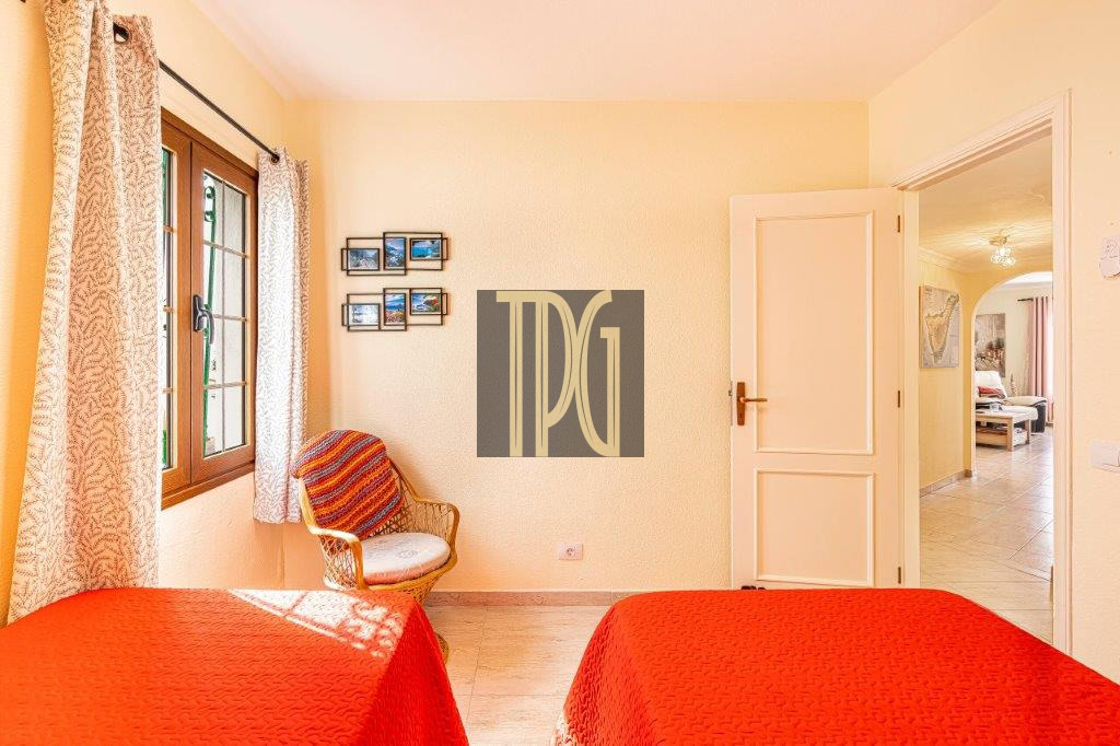 Sunlit bedroom with two red-covered beds, a wicker chair by a window, and an open door to an adjoining room.