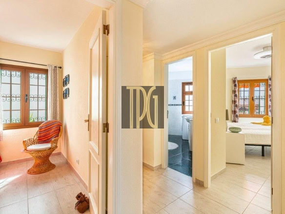 Bright hallway with two open doors leading to a bedroom and a bathroom, plus a wicker chair by a window with patterned curtains.