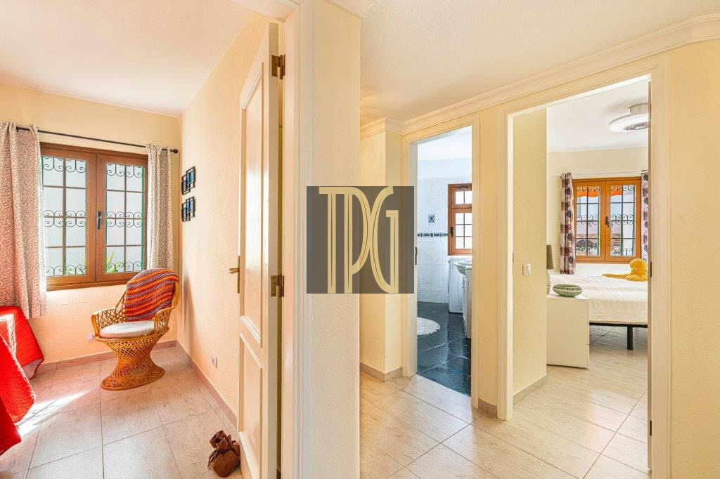 Bright hallway with two open doors leading to a bedroom and a bathroom, plus a wicker chair by a window with patterned curtains.