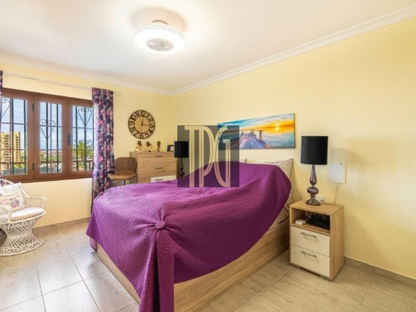 Bright bedroom with a wooden bed, purple quilt, and matching nightstands on yellow walls; window with bars and floral curtains to the left.