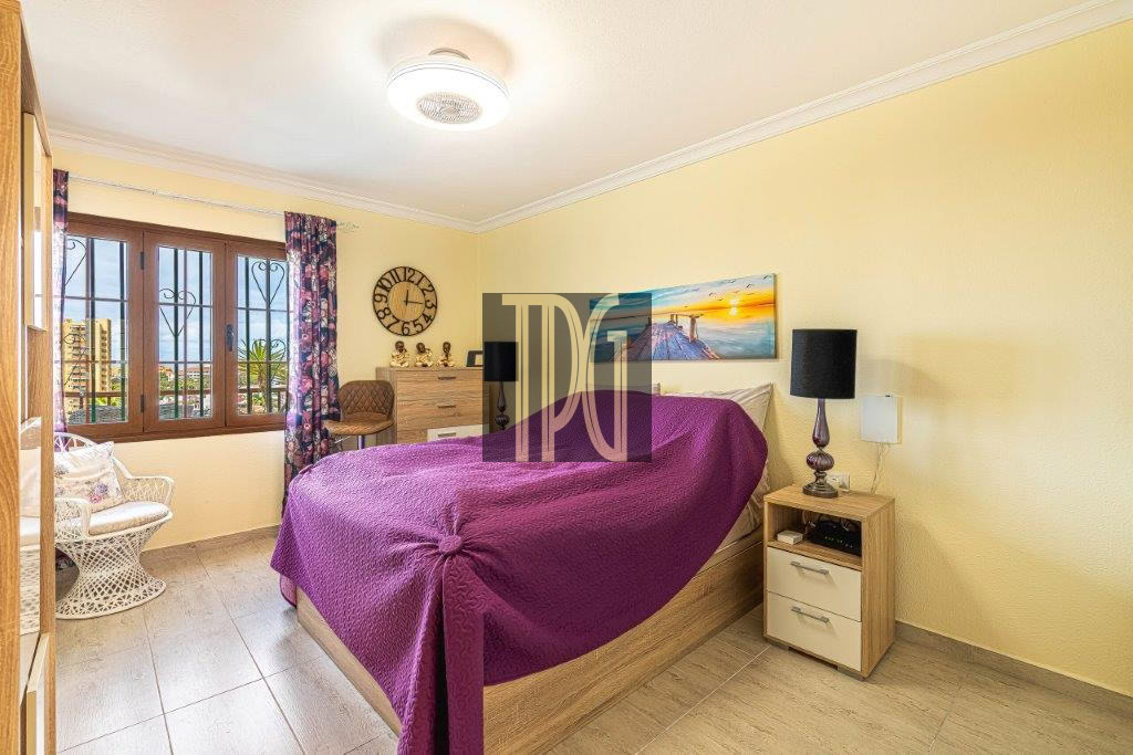 Bright bedroom with a wooden bed, purple quilt, and matching nightstands on yellow walls; window with bars and floral curtains to the left.