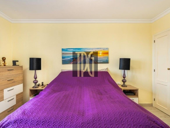 Bedroom with yellow walls and a purple quilted bedspread, two nightstands with black lamps, and a landscape painting above the bed.