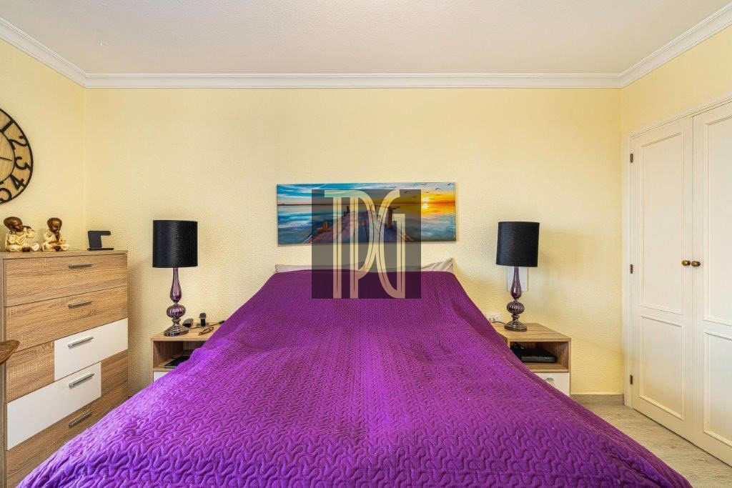 Bedroom with yellow walls and a purple quilted bedspread, two nightstands with black lamps, and a landscape painting above the bed.