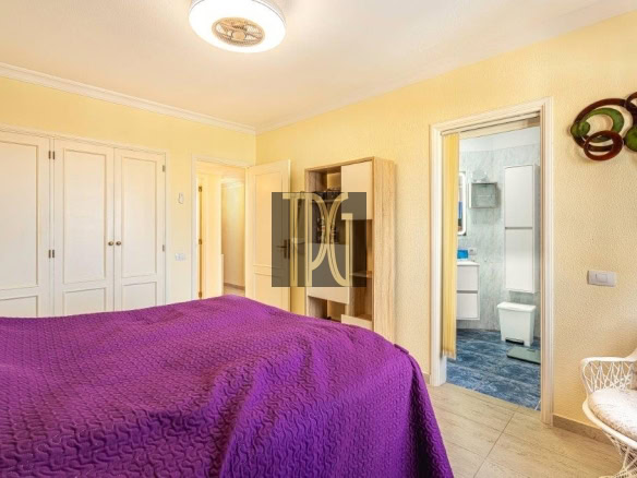 Bright yellow bedroom with a purple quilted bed, white built-in wardrobes along the left, a doorway to a blue-tiled bathroom on the right, and a decorative wall sculpture.