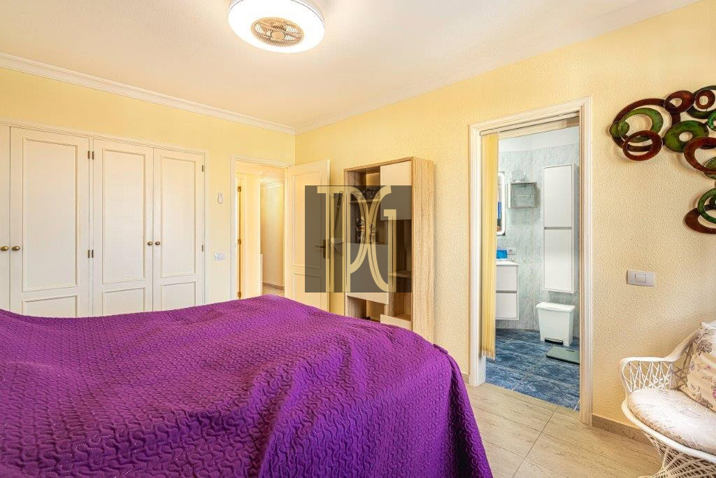 Bright yellow bedroom with a purple quilted bed, white built-in wardrobes along the left, a doorway to a blue-tiled bathroom on the right, and a decorative wall sculpture.