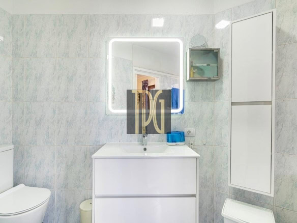 Bright bathroom with a white vanity and backlit square mirror, light blue tiled walls, and a toilet to the left. A tall white cabinet is on the right wall.