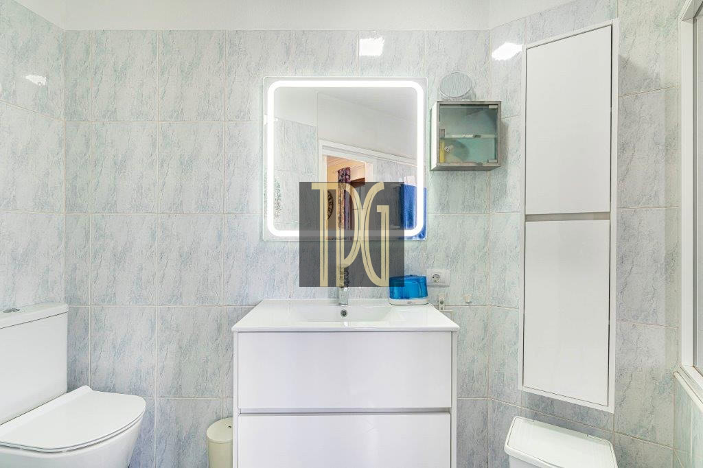 Bright bathroom with a white vanity and backlit square mirror, light blue tiled walls, and a toilet to the left. A tall white cabinet is on the right wall.
