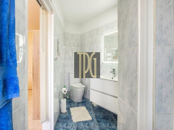 Small blue-tiled bathroom with a white vanity, toilet, and a fluffy rug on dark blue floor tiles, blue towels on the left.