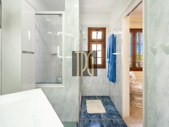 Bright blue-tiled bathroom with a window, blue towel on a rack, and a doorway to a sunlit adjacent room with a wicker chair