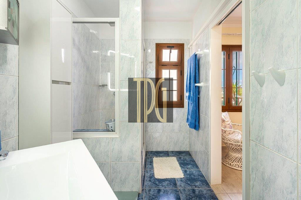 Bright blue-tiled bathroom with a window, blue towel on a rack, and a doorway to a sunlit adjacent room with a wicker chair