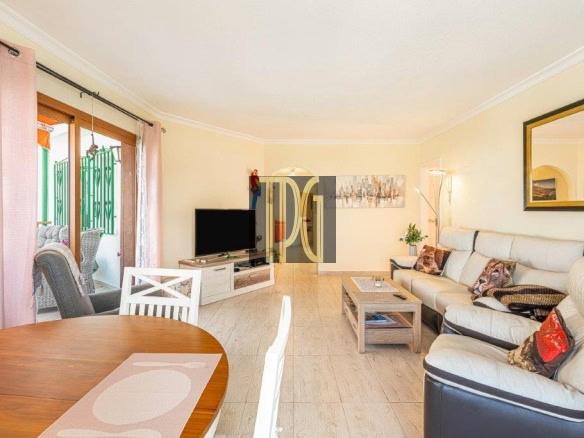 Bright living room with a beige leather sectional, coffee table, and a TV on a low stand; sliding glass doors with pink curtains lead to a balcony outside.