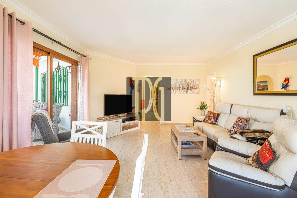 Bright living room with a beige leather sectional, coffee table, and a TV on a low stand; sliding glass doors with pink curtains lead to a balcony outside.